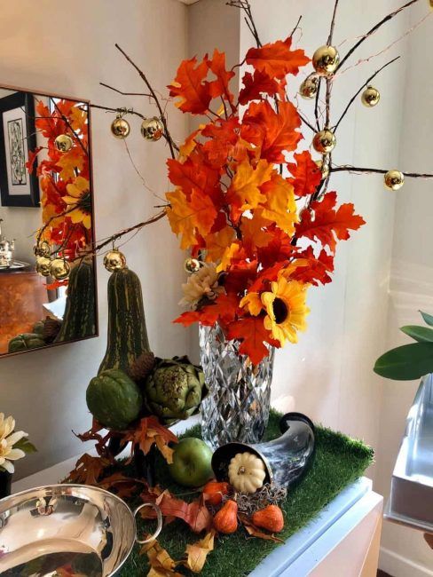 Hojas secas en un florero con algunas bolitas doradas acompañadas de pequeñas calabazas, velas y frutas en un sideboard, decoran una esquina para celebrar Halloween.